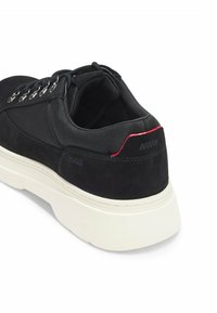 Mustat mokkanahkakengät valkoisella kumipohjalla, joissa on hopeiset nauhakujaan, mustat nauhat ja punainen yksityiskohta kantapäässä. Brändilogo näkyvissä.