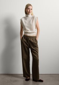 Haut à col roulé sans manches de couleur crème associé à un pantalon large olive avec une taille élastique. La mannequin se tient devant un fond uni.