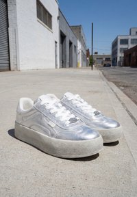 Paire de baskets compensées métalliques argentées avec des lacets blancs placées sur un trottoir en béton urbain sous un ciel clair.