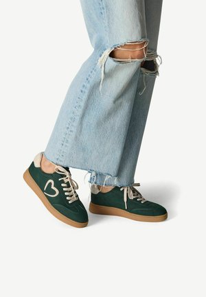 Baskets en suède vert avec des lacets beiges et un logo en forme de cœur sur le côté, associées à un jean large déchiré bleu clair présentant des bords effilochés.