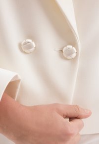 Biała, dopasowana marynarka z teksturowanej tkaniny, z dwoma dekoracyjnymi skręconymi guzikami z przodu, ręka spoczywająca na klapie marynarki.