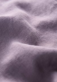 Jasnoliliowy, teksturowany materiał delikatnie się układa, ukazując finezyjny splot oraz gładką powierzchnię z subtelnymi fałdami i cieniami.