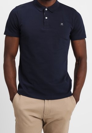 Homme portant un polo bleu marine à manches courtes avec deux boutons et un pantalon beige, debout les mains dans les poches.