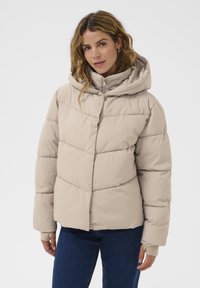 Vaalean beige puffer-takki hupulla, jossa on tikattu pinta, vetoketjusuljenta ja resorit. Päällystetty sinisillä farkuilla.