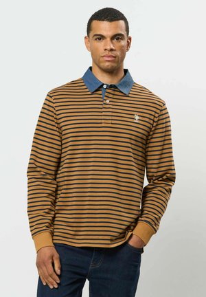 Mann trägt ein langärmeliges Polohemd mit senffarbenen und schwarzen Streifen, blauem Kragen und dunkelblauen Jeans, steht mit einer Hand in der Tasche.