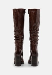 Brązowe skórzane kozaki sięgające za kolano, z marszczeniami, bocznymi zamkami błyskawicznymi i grubymi czarnymi obcasami. Gładka tekstura ogółem.