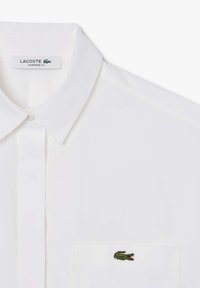 Camicia bianca oversize con colletto classico, chiusura frontale a bottoni e un logo coccodrillo verde ricamato sulla tasca del petto.