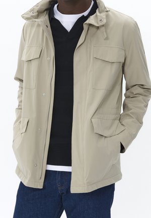 Uomo che indossa giacca beige con tasche con patta sopra maglione nero e camicia bianca, abbinata a jeans blu, mani nelle tasche della giacca.