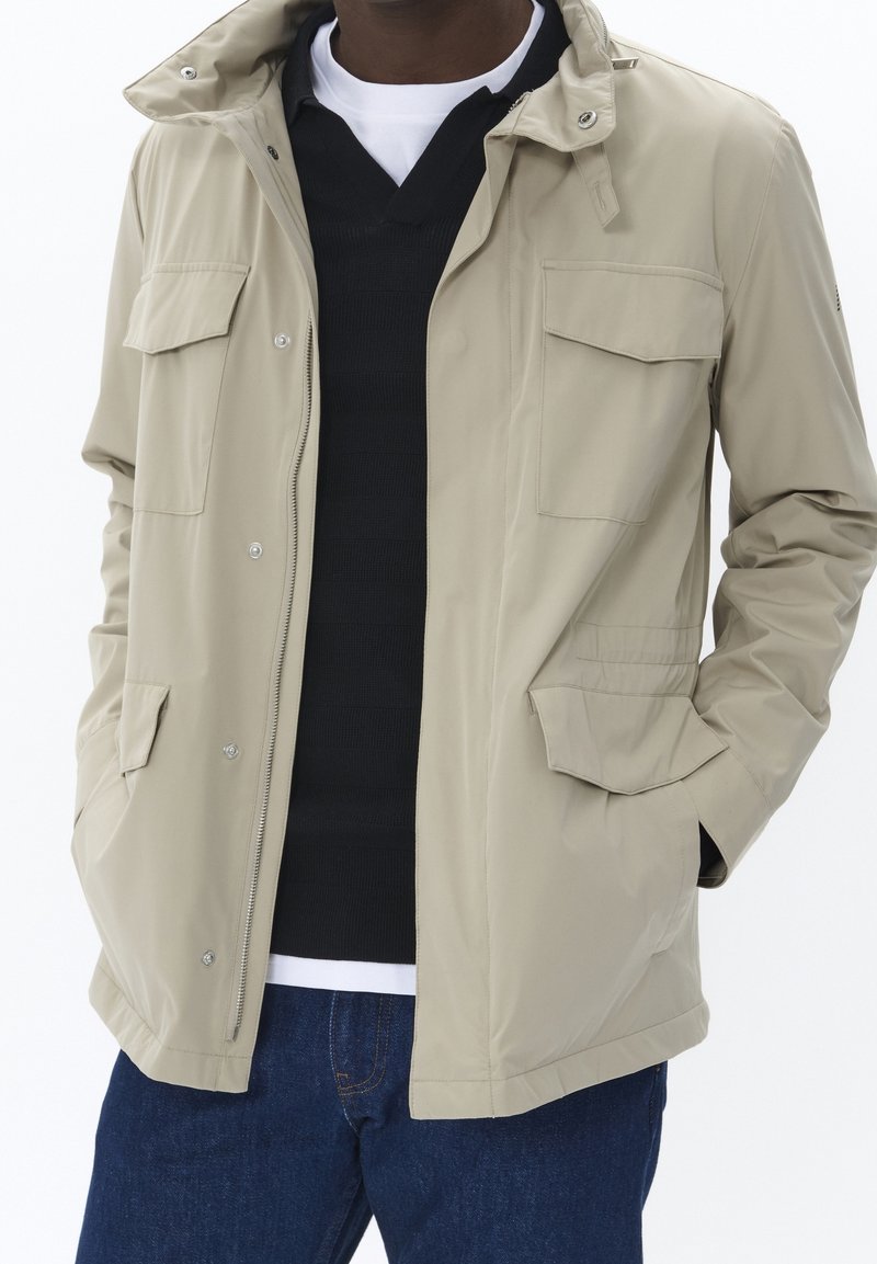 Homme portant une veste beige avec poches à rabat sur un pull noir et une chemise blanche, assortie à un jean bleu, les mains dans les poches de la veste.