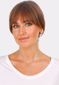 Sølv halskæde med et rundt vedhæng, der har små klare sten, båret over en hvid skjorte. Vedhænget er centreret på kravebenet.