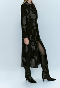 Abito nero a metà lunghezza con un design a fantasia caratterizzato da righe e forme, maniche lunghe e una spacco laterale, abbinato a stivali neri al ginocchio.