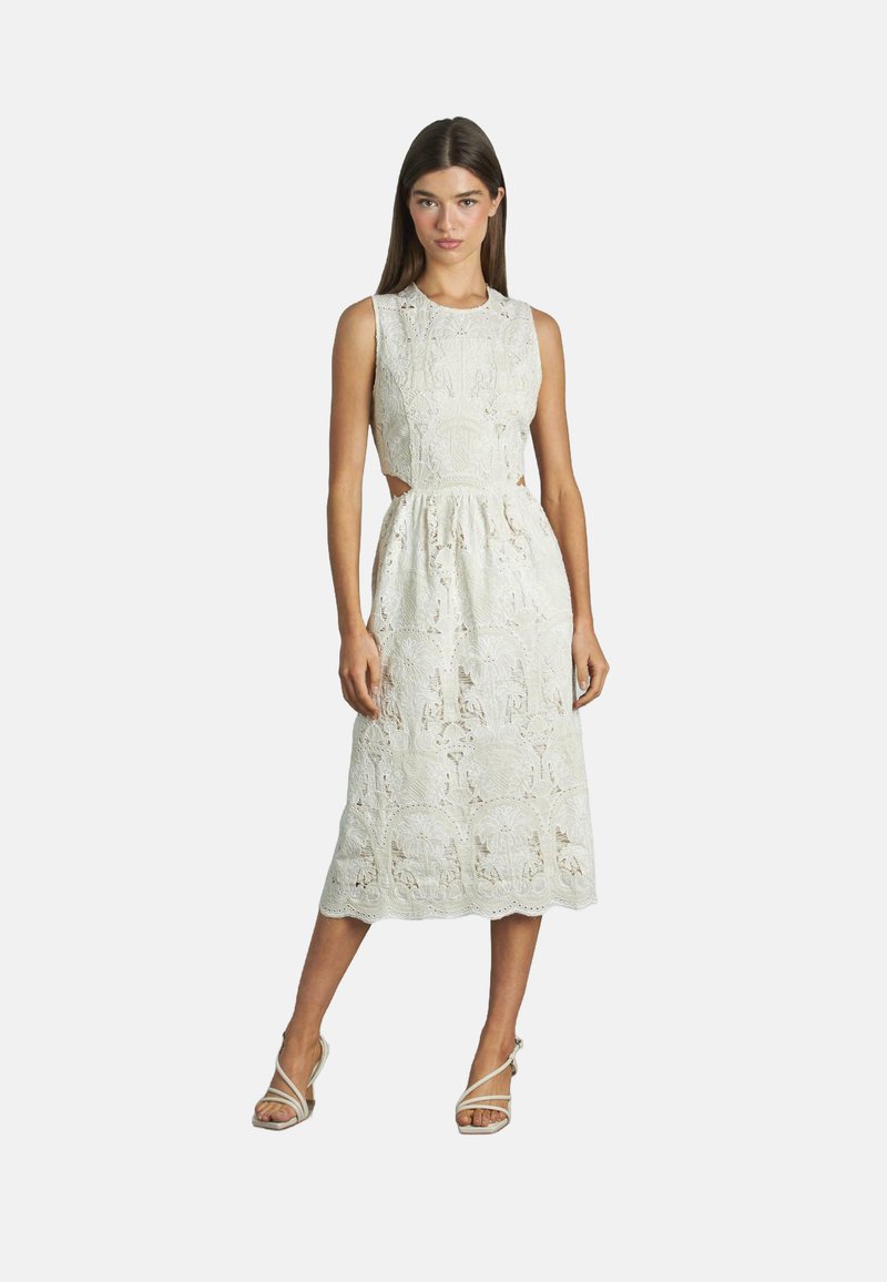 Kleid aus Spitze in Creme mit einem taillierten Oberteil, ausgeschnittenen Seiten und einem knielangen Rock mit aufwendigen Blumenmustern. Riemchensandalen.