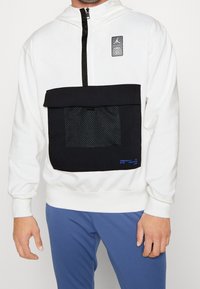 Sudadera blanca con capucha que presenta un bolsillo frontal negro con panel de malla, cierre con cremallera y parche del logo de Jordan. Detalle en texto azul en el bolsillo.