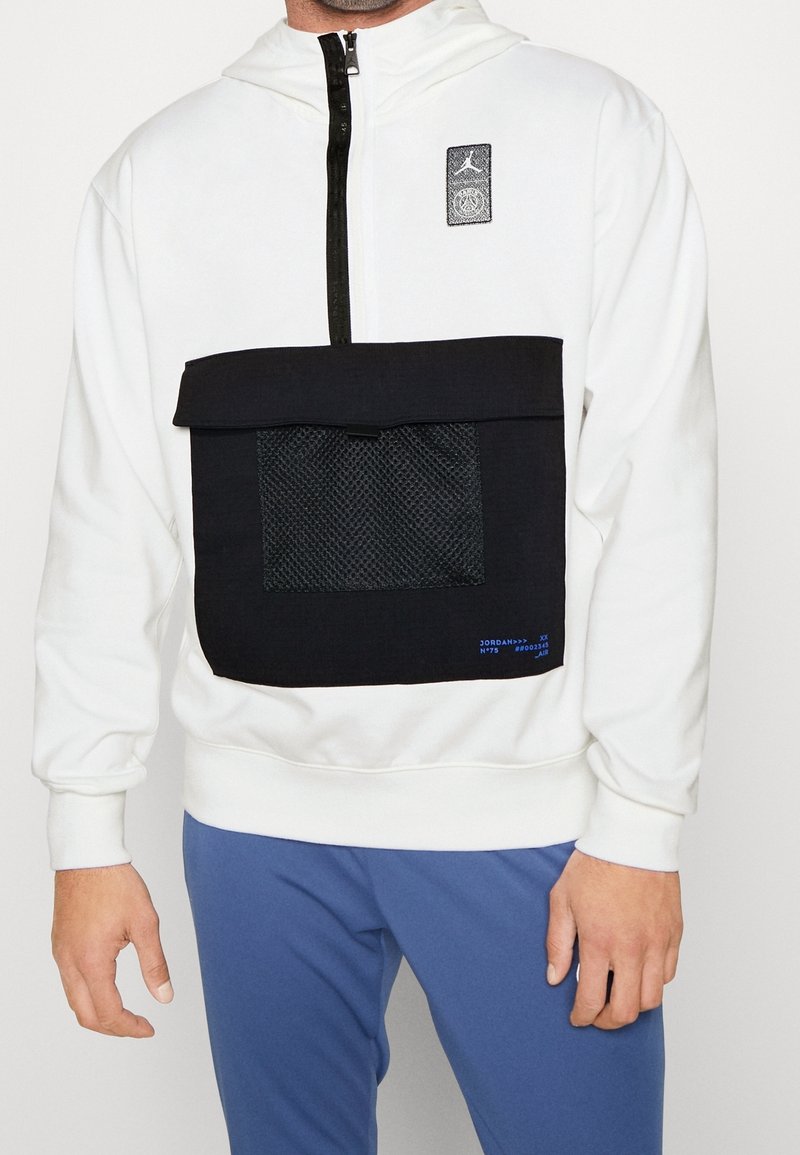 Weißer Kapuzen-Sweatshirt mit einer schwarzen Fronttasche mit Mesh-Panel, Reißverschluss und Jordan-Logo-Patch. Blaue Textdetails auf der Tasche.
