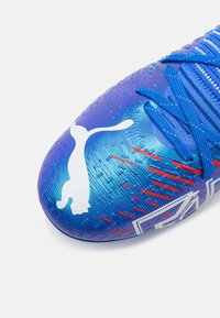 Scarpa da calcio sintetica blu con superficie testurizzata, logo Puma bianco prominente e accenti rossi. Presenta lacci blu e un design elegante.