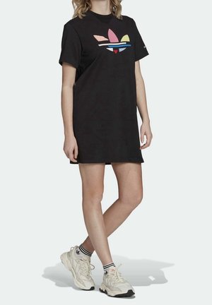 Woman wearing a black short-sleeve dress with a colorful Adidas logo and white Adidas sneakers with striped ankle socks, standing.