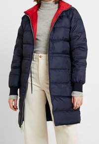 Abrigo acolchado azul marino con forro rojo, cierre de cremallera en el frente, puños elásticos y una silueta larga. Llevado sobre un suéter de cuello alto gris y pantalones blancos.