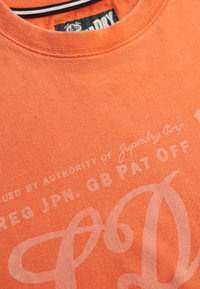 Orange cotton t-shirt featuring a crew neck, faded print with text, and subtle texture. Label shows "Superdry Corp" branding.
