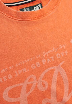 Maglietta in cotone arancione con scollo a girocollo, stampa sbiadita con testo e texture sottile. L'etichetta riporta il marchio "Superdry Corp".