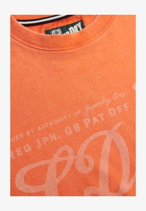 Maglietta in cotone arancione con scollo a girocollo, stampa sbiadita con testo e texture sottile. L'etichetta riporta il marchio "Superdry Corp".