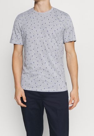 Homme portant une chemise à manches courtes gris clair avec un petit imprimé cactus bleu foncé et un pantalon foncé sur fond uni.