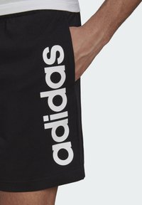Pantalones cortos negros de tela suave, con un logo blanco impreso verticalmente a lo largo del lateral. Una mano está metida en el bolsillo.