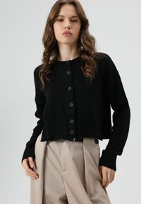 Zwart cropped vest met geribbelde manchetten en zoom, voorzien van zes ronde zwarte knopen aan de voorkant. Model draagt beige hoge taille broek.