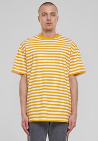 Junger Mann mit kurzen Haaren, der ein gelb-weiß gestreiftes T-Shirt und graue Hosen vor einem schlichten grauen Hintergrund trägt.