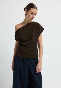 Top asimmetrico marrone realizzato in tessuto morbido, con un design drappeggiato e una vestibilità ampia, abbinato a pantaloni larghi blu Navy.