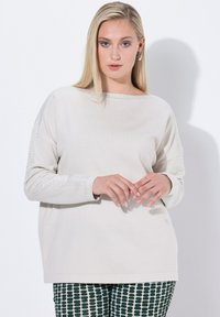 Blonde Frau mit leichter Schminke trägt einen beigen Pullover mit kleinen Löchern an den Ärmeln und gemusterte schwarz-weiße Hosen und steht vor einer weißen Wand.