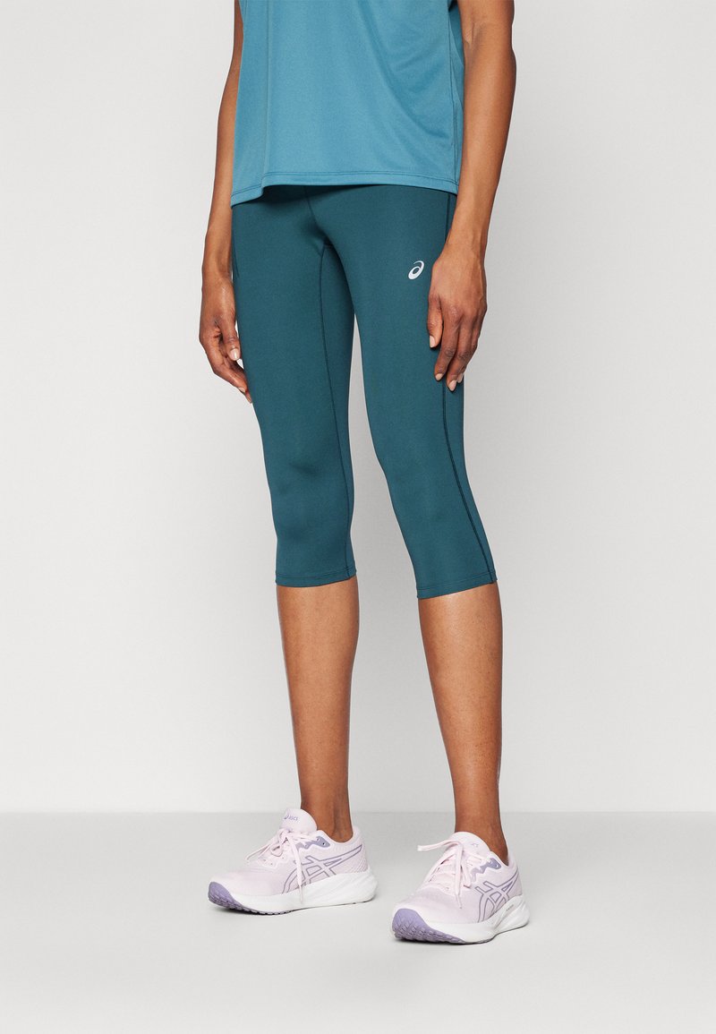 Leggings sportive di colore teal e una maglietta blu, con cuciture curve. Abbinati a scarpe da corsa rosa chiaro con accenti viola.