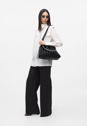 Femeie purtând pantaloni negri largi și o cămașă albă, ținând o geantă neagră matlasată cu detalii din lanț peste umăr.