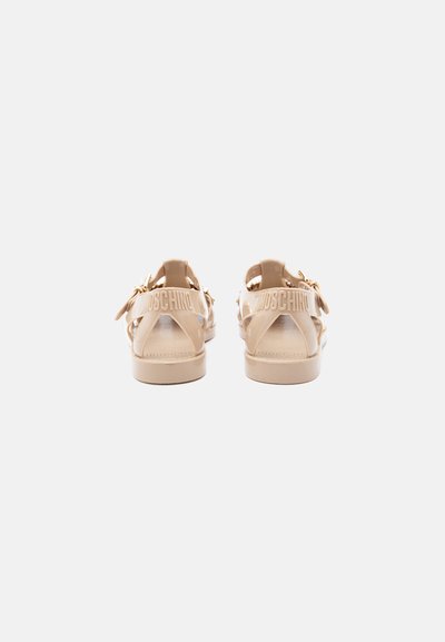 Sandales Moschino beige brillantes avec une bride à boucle, bout ouvert, et nom de la marque embossé sur les brides arrière, vue de dos.