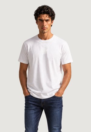 Jeune homme aux cheveux bouclés, debout face à l'avant, les mains dans les poches, portant un t-shirt blanc uni et un jean bleu foncé.