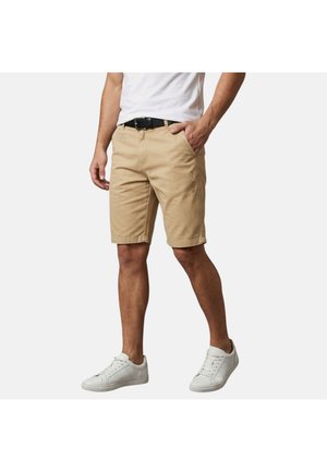 Homme portant un short beige jusqu'au genou, un t-shirt blanc, une ceinture noire et des baskets blanches, debout avec une main dans la poche.