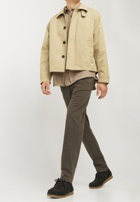 Beżowa wodoodporna kurtka z czarnymi guzikami, pod nią brązowa koszula w kratę, oliwkowe spodnie i czarne buty z brązowymi podeszwami.