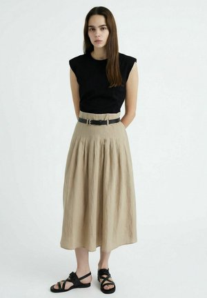 Frau mit langen braunen Haaren, die ein schwarzes ärmelloses Oberteil, einen beigefarbenen plissierten Midirock mit schwarzem Gürtel und schwarze Sandalen trägt, vor weißem Hintergrund stehend.