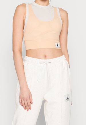 Woman wearing light peach mesh sports bra over white tank top and loose white sweatpants with drawstring, standing against plain background.