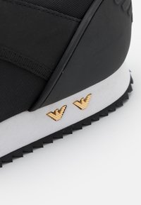 Černá sneaker s texturovaným látkovým svrškem, kontrastní bílou podešví a zlatými křídly tvarovanými logy na boku. Gumová podešev se zigzagovým vzorem.