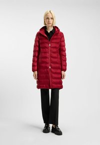 Röd vadderad jacka med huva, med vertikal sömnad, en hel dragkedja och sidofickor. Kombinerad med svarta randiga byxor och skor.