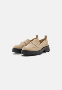 Beige zamšaini loafers ar melnu masīvu zoli, ar plakanu dizainu, elastīgām sānu daļām un dekoratīvu jostu priekšpusē.