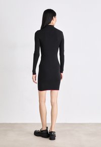 Rochie neagră din tricot cu nervuri, cu mâneci lungi, o siluetă ajustată și un tiv roșu. Asortată cu încălțăminte groasă neagră.