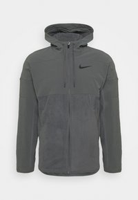 Felpa grigia con zip e tasca anteriore, con una parte superiore liscia e una parte inferiore testurizzata. Include un cappuccio e un logo nero.