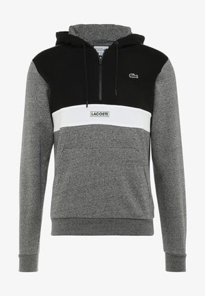 Sweat à capuche gris et noir avec une bande horizontale blanche, une poche avant, une fermeture éclair au niveau du col et le logo Lacoste sur la poitrine et la bande.