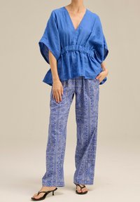 Niebieska lniana bluzka z dekoltem w kształcie litery V i marszczonym pasem, połączona z luźnymi, wzorzystymi niebieskimi spodniami z misternymi wzorami. Czarne klapki.