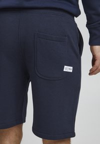 Pantaloni scurți din bumbac bleumarin, cu un buzunar posterior, textură netedă și tiv cusut. Include un mic eticheta de marcă pe buzunar.