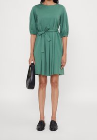 Frau, die ein knielanges grünes Kleid mit Puffärmeln und einem Taillengürtel trägt, eine schwarze Handtasche hält und schwarze Slipper anhat.