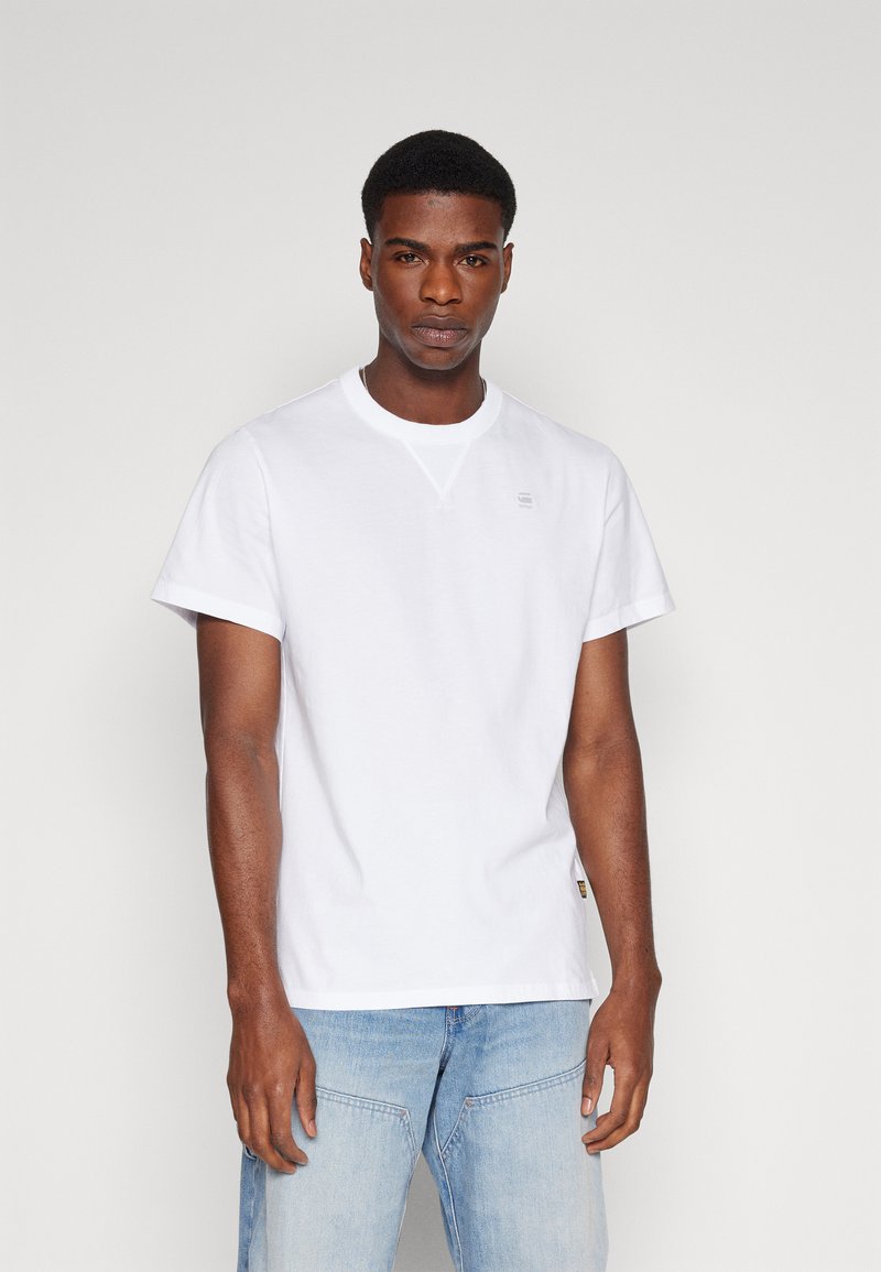 T-shirt blanc à manches courtes en coton, avec un col rond, un logo discret sur la poitrine et une coupe décontractée. Associé à un jean bleu clair.