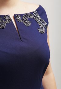 Námořnické šaty s pevně strukturovaným výstřihem, zdobené vyšívanými korálky v točeném vzoru. Měkká, texturovaná látka s mírným leskem.