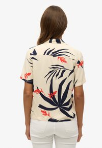 Camisa de manga corta en crema con patrón de hojas tropicales en azul marino y rojo, que presenta un corte relajado y cuello clásico, hecha de tela ligera.